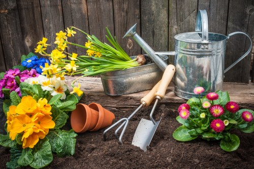 The image depicts a gardening scene in a small outdoor garden area, featuring a section of dark, freshly turned soil in the foreground with various gardening tools and decorative potted plants arranged on the surface. There are two terracotta plant pots, one resting on its side and the other upright, alongside a small garden fork and trowel with wooden handles, positioned across a galvanized watering can with a curved handle and a spout. A rectangular metal container holds a bunch of yellow daffodils with long green stems, while a cluster of vibrant pink and yellow flowering plants with lush green leaves is situated nearby. The background consists of a weathered wooden fence, framing the scene and providing a rustic backdrop. The lighting is natural, suggesting a bright day with soft shadows. This garden setup emphasizes typical elements of outdoor maintenance and planting, aligning with services offered by Gardening Services Norwood in the Norwood area, South London, and reflects an inviting, well-organized garden environment suitable for landscaping, lawn care, and plant care activities.