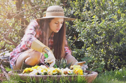 A young woman with long brown hair, wearing a straw hat, a red and white checkered shirt, and gardening gloves, is kneeling on a well-maintained lawn in a garden. She is using her hands to tend to a flower bed that contains a variety of blooming flowers in shades of yellow, white, and purple. The garden is lush with green foliage, including dense bushes and trees in the background, creating a natural and vibrant outdoor space. The sunlight filters through the leaves, illuminating the scene with warm, natural light, suggesting a clear, pleasant day suitable for gardening. This setting showcases typical features of local gardens in Norwood, incorporating neatly bordered flower beds and healthy grass, highlighting professional gardening and landscape maintenance services provided by Gardening Services Norwood in the area.