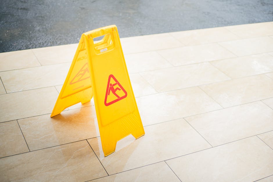 A yellow caution wet floor sign with red warning symbols is placed on a beige tiled patio or entrance area, indicating a potentially slippery surface. The paving stones are textured and arranged in a 