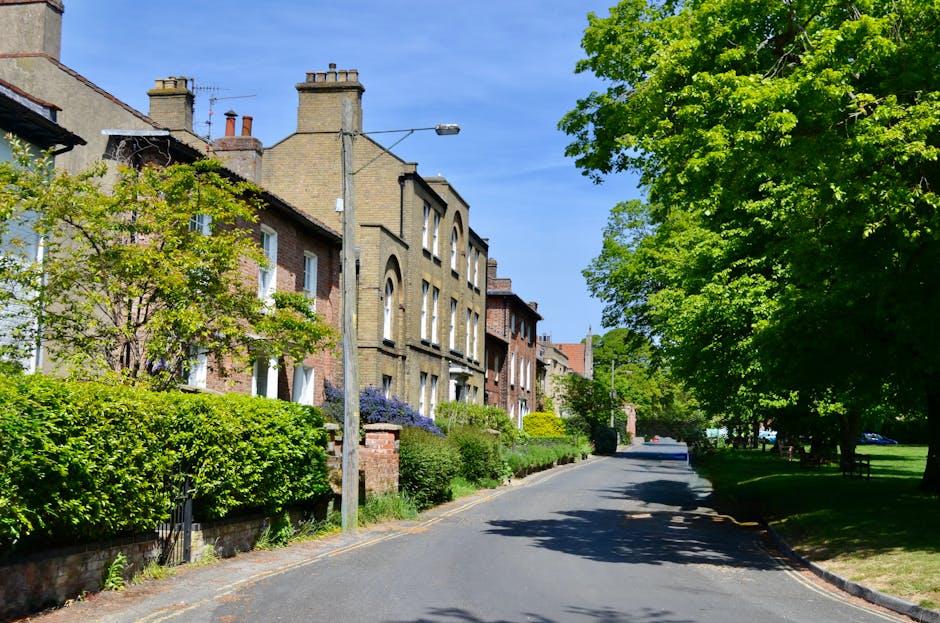 A quiet residential street featuring Victorian terraced houses with brick facades and tall chimneys, lined with neatly maintained front gardens filled with lush green shrubs, small flowering trees, an